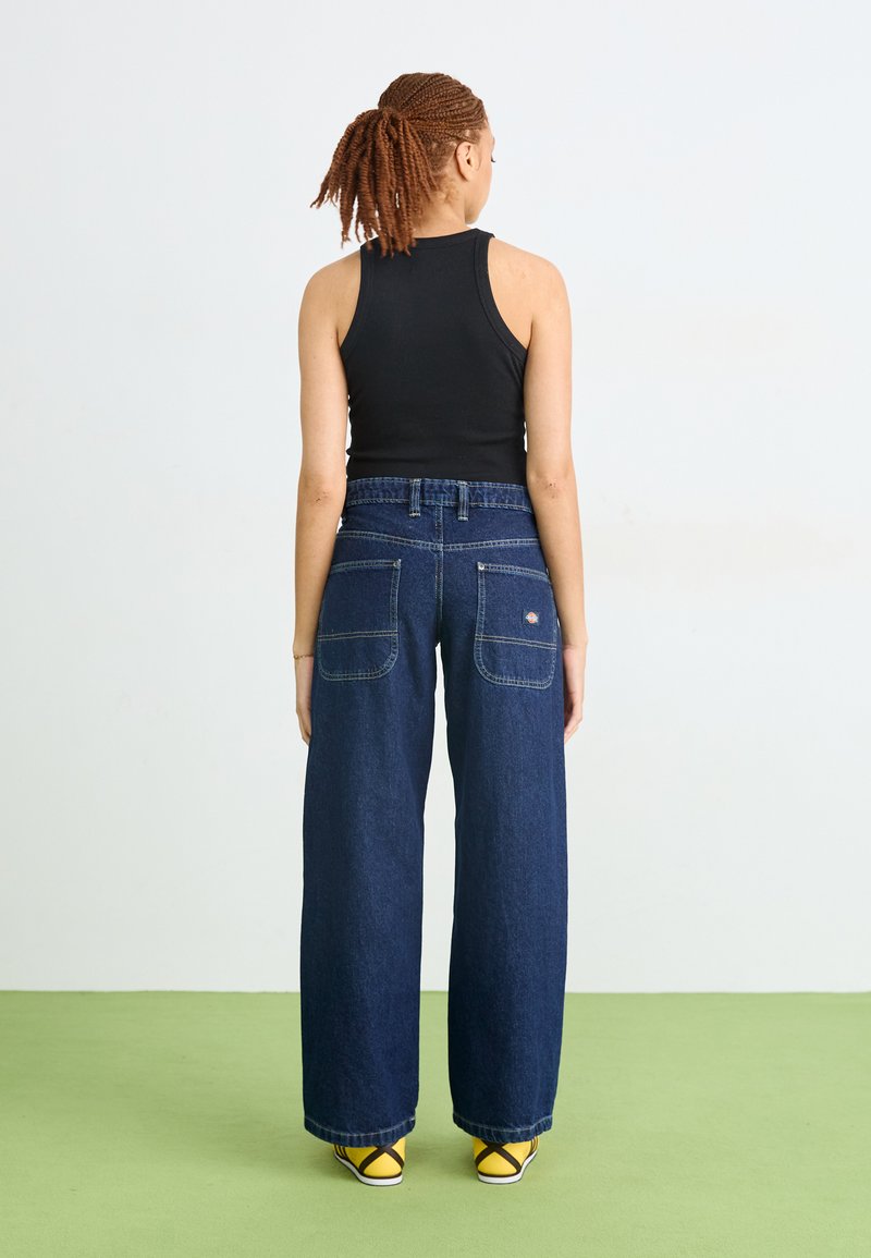 Jeans in denim a gamba larga di colore blu scuro con vita alta, dotati di doppie tasche posteriori e cuciture a contrasto. Abbinati a scarpe gialle.