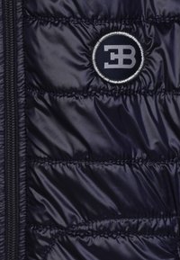 Giacca imbottita blu navy con chiusura a zip, caratterizzata da una patch rotonda con logo in argento e blu scuro sul lato sinistro. Tessuto morbido e lucido.