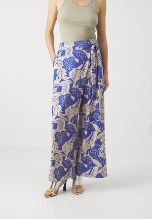 La femme porte un pantalon large bleu et beige avec un grand motif de feuilles, un haut sans manches beige, et des sandales à talons hauts argentées.