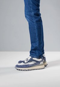 Siniset ja harmaat urheilukengät, jotka on valmistettu mokkanahasta ja verkkokankaasta, paksulla kumipohjalla ja valkoisilla nauhoilla.