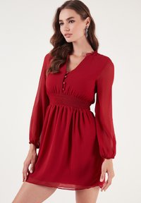 Rode chiffon jurk met lange mouwen, V-hals, knoopdetails, geschubde taille en uitlopende rok. De doorschijnende textuur voegt zachtheid toe.