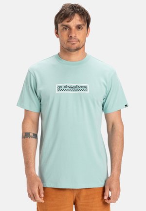Lys turkis bomulds T-shirt med et grafisk logo med ternede detaljer i midten. Korte ærmer og design med klassisk rund hals.