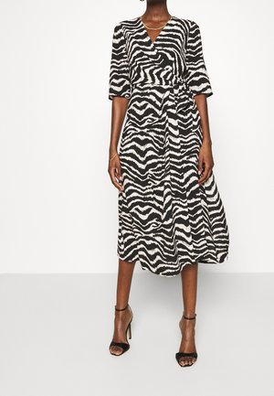 Femme portant une robe portefeuille à imprimé zèbre noir et blanc avec des manches trois-quarts et des sandales noires à talons hauts.