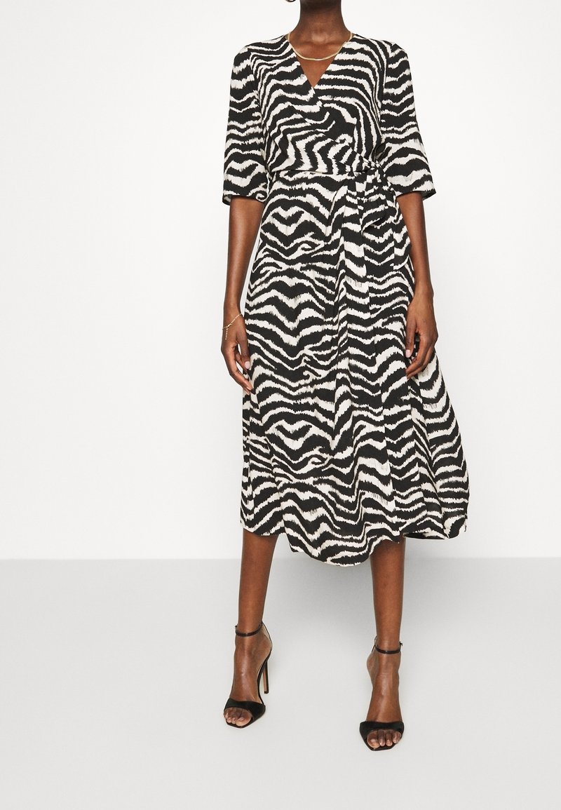 Femme portant une robe portefeuille à imprimé zèbre noir et blanc avec des manches trois-quarts et des sandales noires à talons hauts.