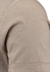 Top de algodón beige con una textura suave, que presenta una costura de hombro curvada y un acento de costura roja en el puño.