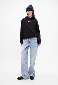 Czarna bluza z kapturem, z kieszenią z przodu, luźny krój; zestawiona z jasnoniebieskimi spodniami typu wide-leg i czarnymi sneakersami. Model nosi ciemną czapkę beanie.