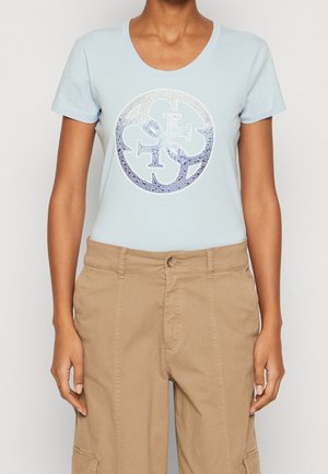 Vrouw met een lichtblauwe T-shirt met korte mouwen en een ronde halslijn, voorzien van een glanzend cirkelvormig logo, in combinatie met beige cargobroeken.