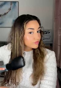 Brosse à cheveux noire avec une tête large et courbée et des poils. Cheveux bruns longs et ondulés en cours de coiffage. Pull blanc texturé avec des embellissements.