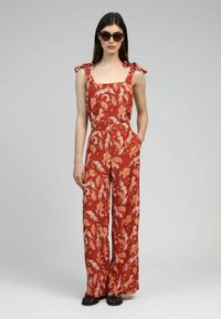 Combinaison florale en rouge rouille avec des motifs blancs et oranges, décolleté carré et bretelles en nœud. Dispose de jambes larges et de poches latérales.