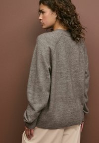 Pulover gri tricotat cu un finisaj texturat, tiv ondulat și un design cu umeri drop, purtat peste pantaloni largi de culoare crem.