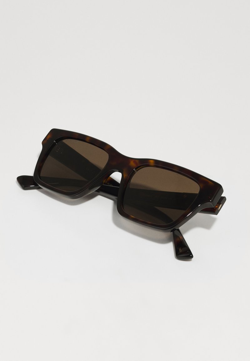 schildpadkleurige zonnebril met vierkante monturen, donkere lenzen en dikke armen. Opvallende uitsparingen op het montuur en subtiele gouden accenten op de schakels.