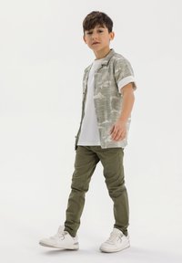 Camicia a maniche corte di colore verde chiaro con motivo e dettagli bianchi, abbinata a una t-shirt bianca e pantaloni verde oliva. Indossata con sneakers bianche.