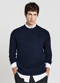 Marineblauer Strickpullover mit Rundhalsausschnitt und langen Ärmeln, über einem weißen Hemd mit sichtbaren Manschetten getragen. Einfaches Design und strukturierter Stoff.