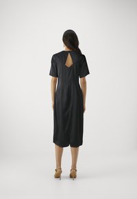 Svart kjole med korte ermer, kne-lengde, med en nøkkelhullsutskjæring bak og glatt satin-finish. Brukt med lysebrune høye sandaler.