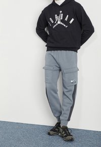 Czarna bluza z kapturem z logo "JORDAN", szare spodnie dresowe cargo z kieszeniami, czarne buty Nike oraz gładka faktura, na tle białej ściany i niebieskiej podłogi.