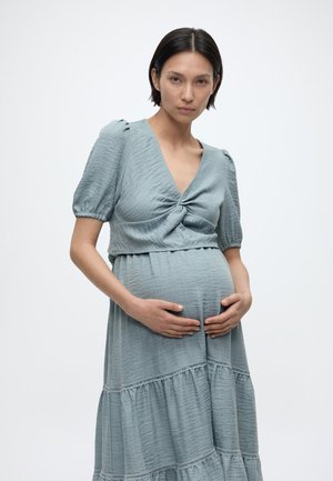 Donna incinta con capelli corti e scuri che indossa un vestito azzurro chiaro con trama, che tiene delicatamente la pancia su uno sfondo bianco uniforme.