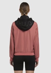 Veste bombardier rose avec une capuche en polaire noire comportant un logo "KHUJO" en relief. Tissu doux, poignets et taille élastiques.