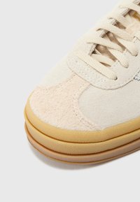 Μπεζ σουέτ αθλητικό παπούτσι με ανοιχτό ροζ λεπτομέρειες και στρογγυλή λαστιχένια σόλα. Διαθέτει επίπεδα κορδόνια και υφή στην επιφάνεια.