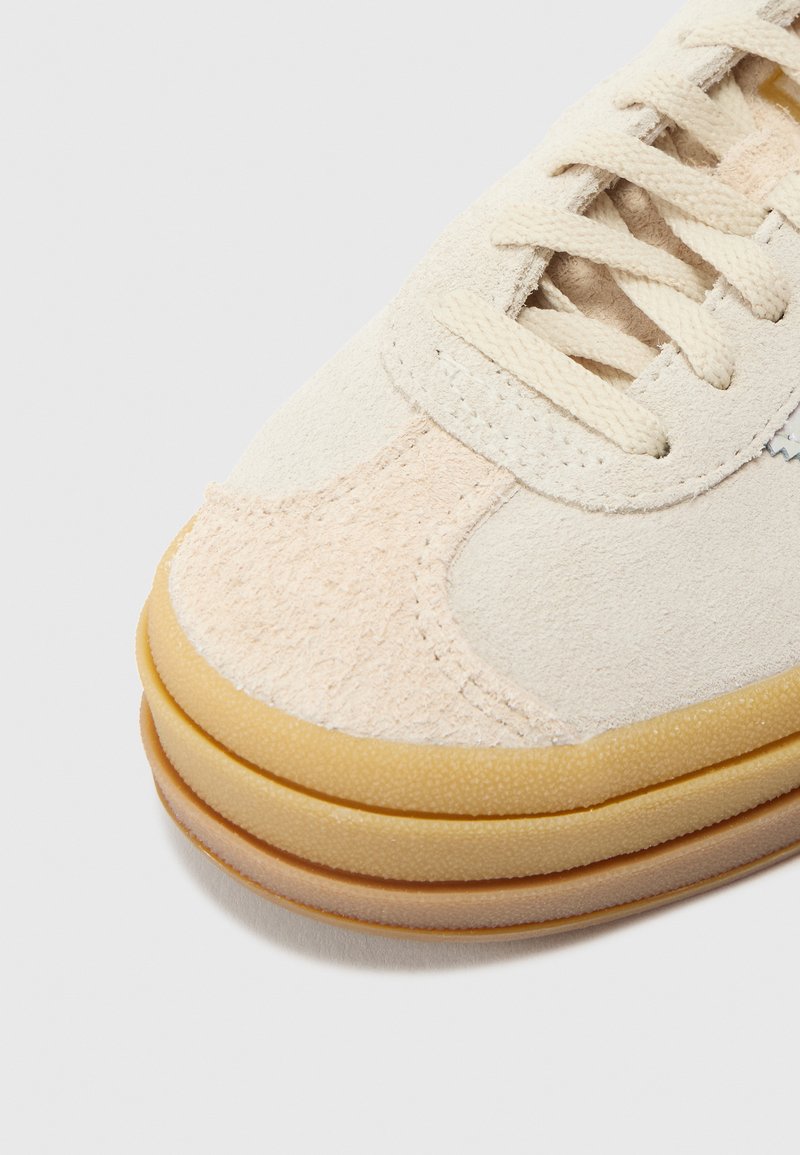 Beige mockasinsneaker med ljusrosa detaljer och rund gummisula. Har platta snören och texturerad yta.