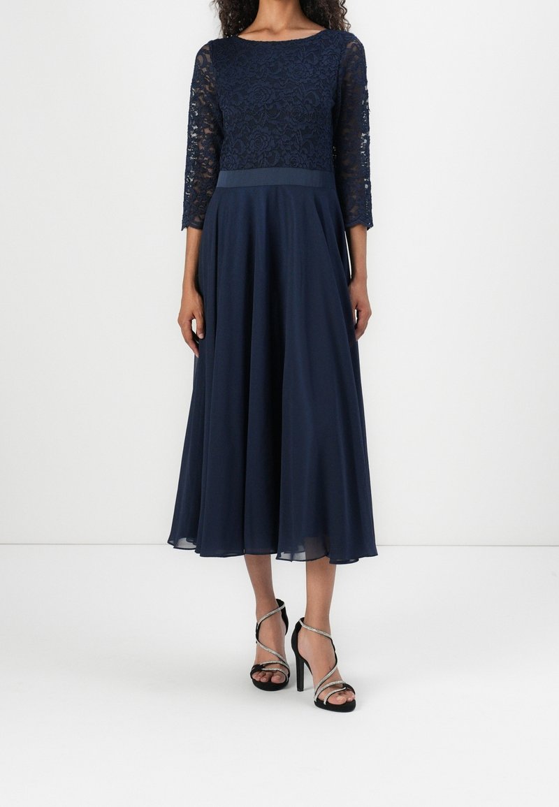 Robe marine avec un corsage en dentelle et des manches trois-quarts, jupe fluide et ceinture en satin. Associée à des talons noirs à lanières.