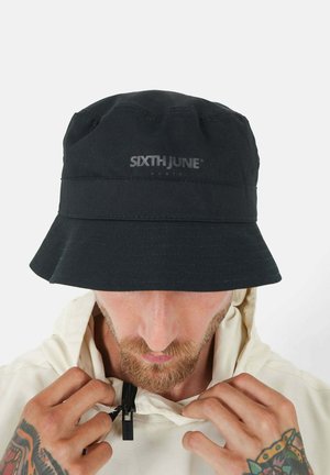 Schwarzer Bucket Hat mit strukturiertem Finish und einem Logo auf der Vorderseite. Worn mit einer hellen Jacke mit Reißverschluss.