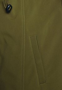 Tissu de veste vert olive avec une texture lisse, doté d'une poche latérale et de détails de coutures visibles. Fermeture par cordon au niveau du col.
