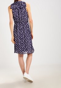 Žena stojí s otočeným zadkem k divákovi, na sobě má fialové šaty se vzorem a volánkovým límcem, a bílé tenisky.