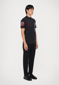T-shirt nero a maniche corte con testo rosso e dettagli a toppa, abbinato a pantaloni scuri e sneakers nere, indossato da un modello maschile.