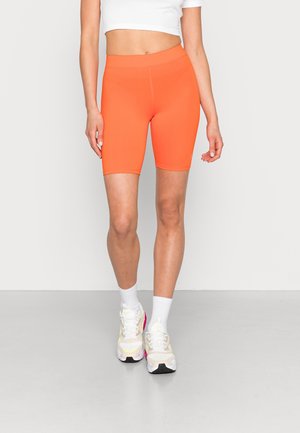 Persoon draagt feloranje aansluitende biker shorts, wit cropped topje, witte sokken en witte sneakers met roze accenten, staand op een grijze vloer.