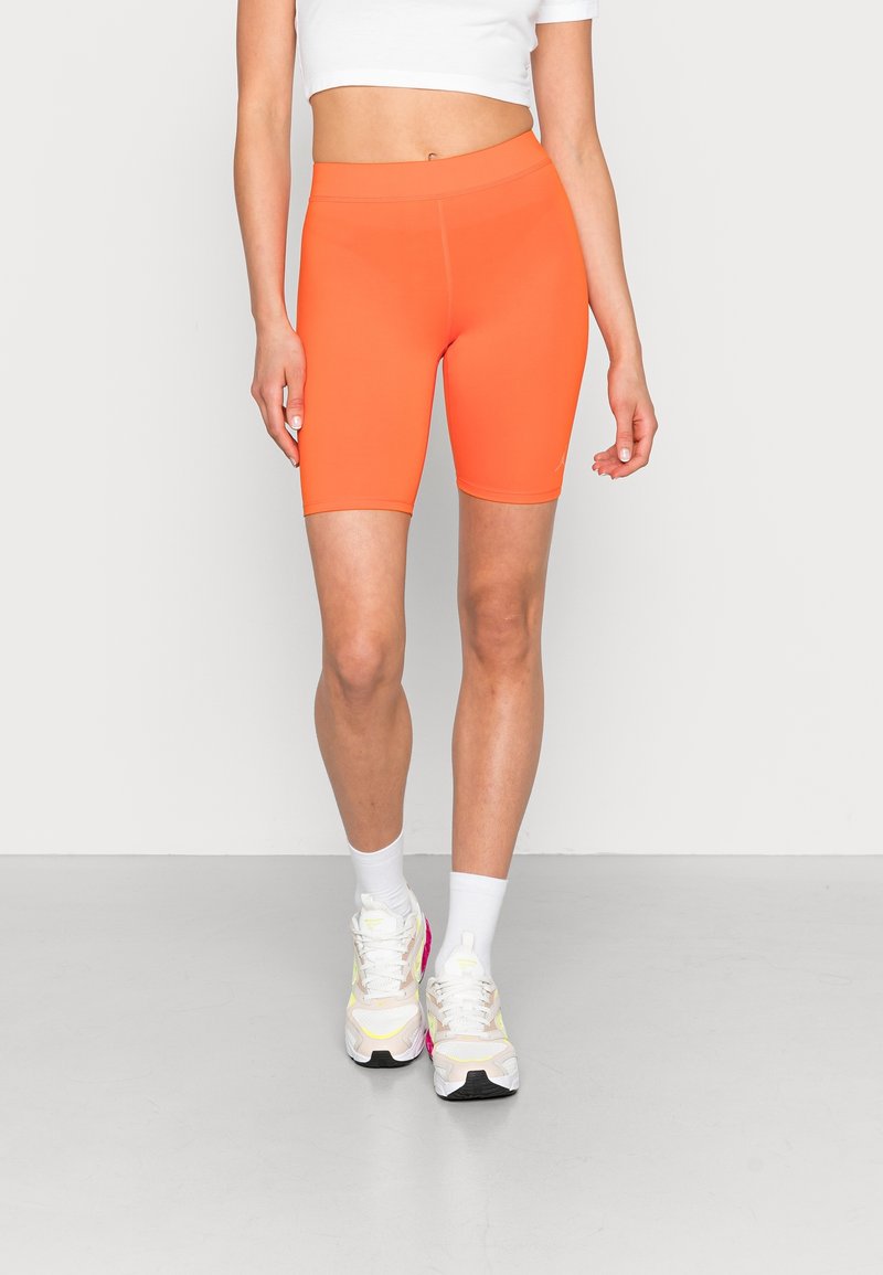 Personne portant un short cycliste moulant orange vif, un top court blanc, des chaussettes blanches et des baskets blanches avec des touches de rose, debout sur un sol gris.