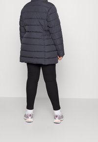 Persona che indossa un giubbotto imbottito scuro, leggings neri, calzini bianchi e scarpe da corsa argentate, in piedi contro uno sfondo chiaro uniforme, vista da dietro.