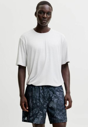 Mand iført en ensfarvet hvid kortærmet t-shirt og mørke mønstrede shorts stående mod en hvid baggrund.
