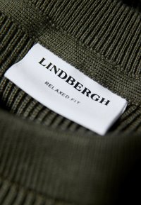 Olivengrüner Strickpullover mit gerippter Textur; verfügt über ein weißes Etikett mit der Aufschrift "LINDBERGH" und "RELAXED FIT" in Schwarz gedruckt.