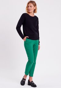 Pull en texture noire à manches longues associé à un pantalon slim-fit vert. La tenue est complétée par des chaussures à talons carrés noires.