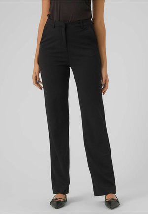 Femme portant un pantalon noir droit, un haut noir et des ballerines noires pointues, debout devant un fond gris uni.