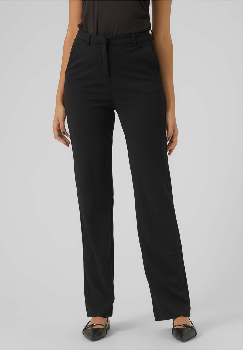 Femme portant un pantalon noir droit, un haut noir et des ballerines noires pointues, debout devant un fond gris uni.