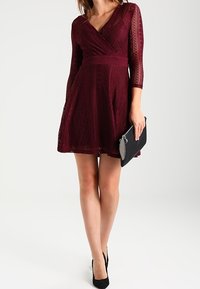 Bordeaux kanten wikkeljurk met driekwart mouwen, A-lijn rok, textuurpatroon en een zwarte clutch met een decoratieve franjes.