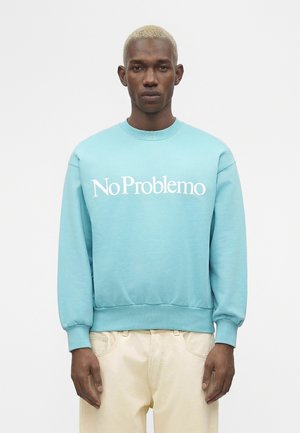 Homme aux cheveux blonds clairs portant un sweat-shirt bleu avec le texte « No Problemo » et un pantalon beige clair, debout devant un fond blanc uni.