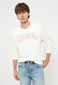 Mladý muž s kučeravými vlasmi, oblečený v bielej mikine s červeným nápisom „DIESEL“ a svetlomodrých džínsach, stojaci pred bielym pozadím.