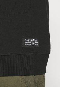 Svart hoodtröja med ribbad nederkant och etikett som har "TOM TAILOR DENIM" i vit text. Tyget verkar slätt med en avslappnad passform.