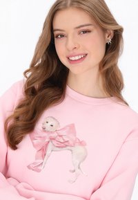 Világos rózsaszín pulóver, amelyen egy nagy rózsaszín masnival díszített kutya grafikája látható. Az anyaga puhanak tűnik, laza szabású és kerek nyakkivágással.