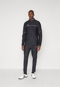 Survêtement de sport noir avec des accents blancs, inclut une veste zippée et un pantalon assorti, fabriqué en matériau lisse et léger. Porté avec des chaussures de sport blanches.