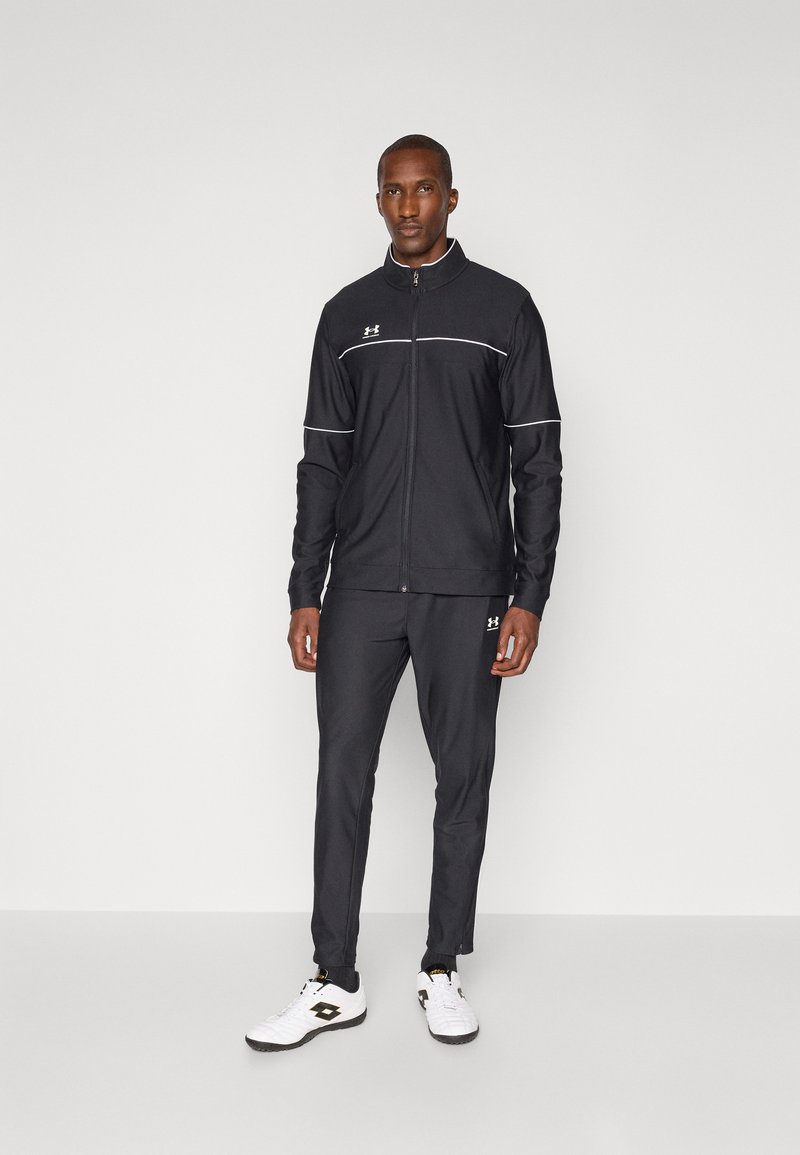 Survêtement de sport noir avec des accents blancs, inclut une veste zippée et un pantalon assorti, fabriqué en matériau lisse et léger. Porté avec des chaussures de sport blanches.