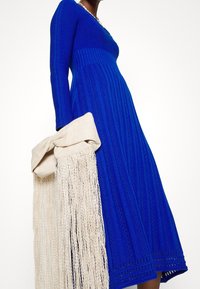 Robe en tricot d'un bleu vibrant, avec une texture côtelée et des manches longues. Accompagnée d'un sac crème orné de franges.
