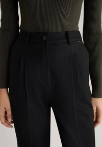 Pantalons noirs ajustés avec une texture lisse, dotés de plis, d'une fermeture à bouton et de poches latérales. Assorti avec un haut côtelé vert olive.