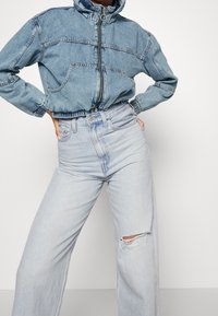 Džínová bunda s vysokým límcem, zapínáním na zip a elastickým lemem, v kombinaci se světle modrými džínami s vysokým pasem a zdobenými roztrženými koleny.