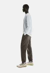 Man standing in profile wearing a white long-sleeve shirt, brown jogger pants, and beige athletic shoes with hands in pockets.