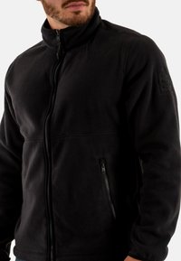 Chaqueta de polar negra con cuello alto, cremallera frontal y bolsillo lateral con cremallera. Presenta una tela texturizada y un logotipo sutil en la manga.