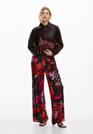 Woman wearing a dark brown leather jacket and vibrant red, pink, and black patterned wide-leg pants, holding a dark red handbag.