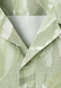 Camisa de color verde claro con un sutil patrón de hojas, que presenta un cierre de botón y una textura suave y mate.